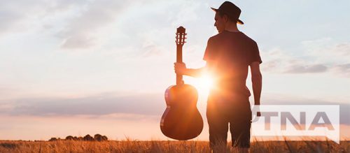 Man holding a guitar in a field against a sunset